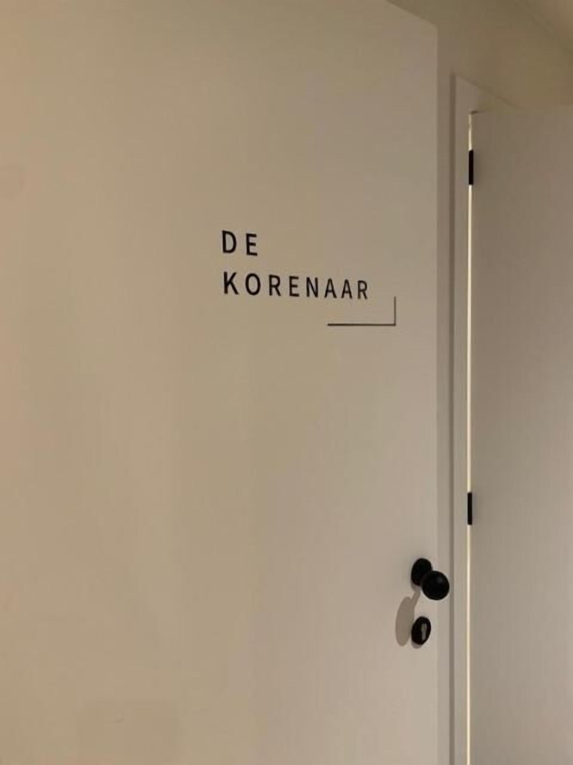 Een witte deur met de tekst “DE KORENAAR” en een dunne horizontale lijn erboven.