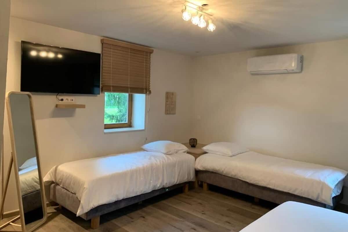 A room with two single beds, a flat-screen TV mounted on the wall, a window with blinds, and an air conditioning unit.