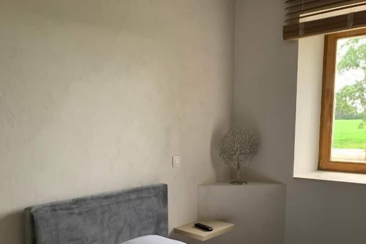 A bedroom with a bed, a nightstand, a wall-mounted lamp, and a window with a view of greenery.