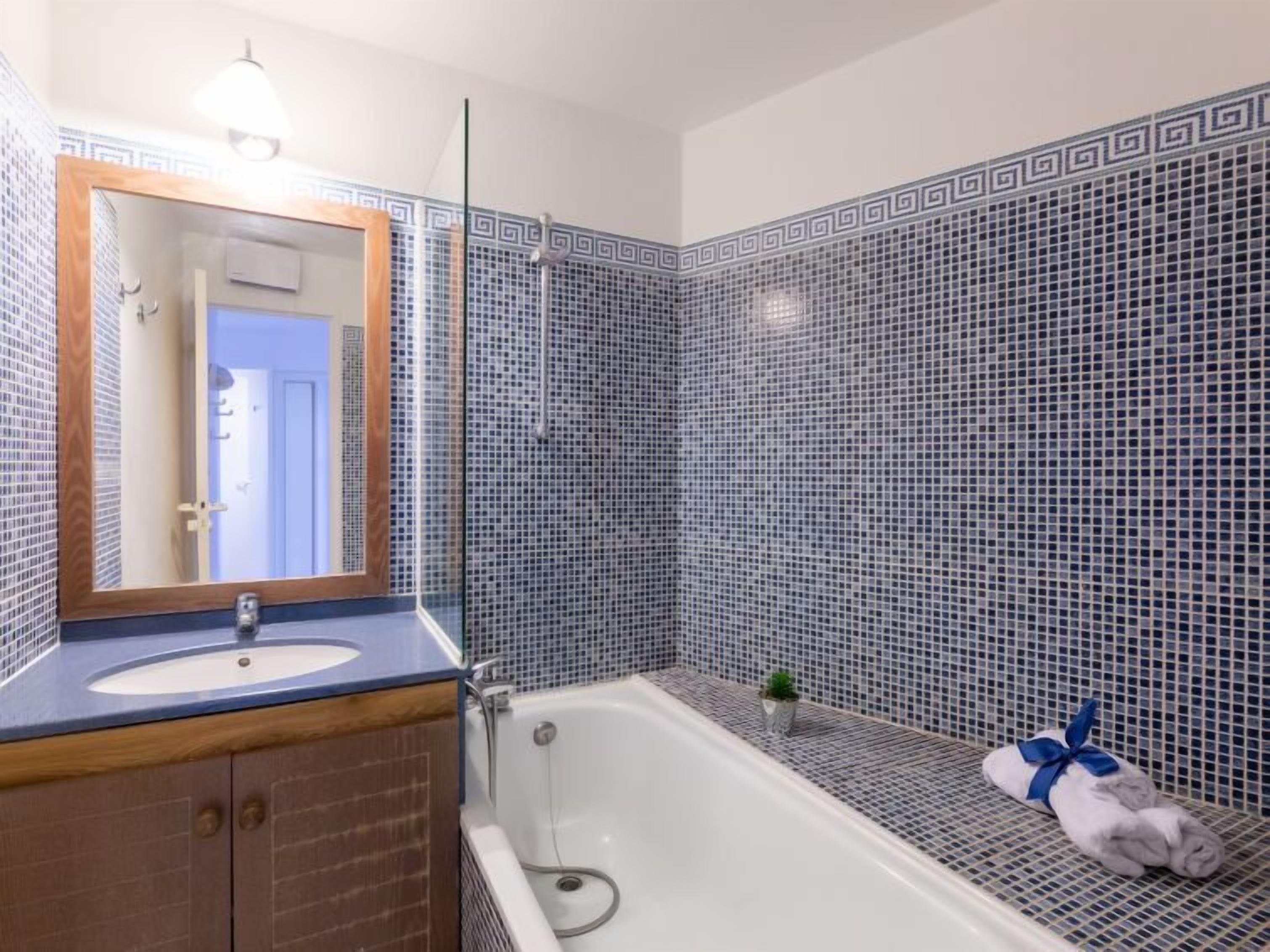 Une salle de bain avec une baignoire blanche, un meuble-lavabo en bois, un miroir et un mur recouvert de carreaux formant un motif géométrique.