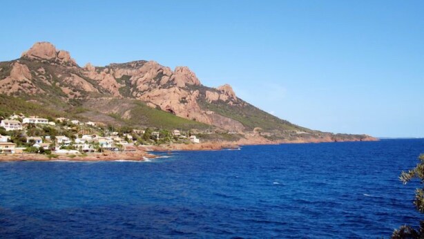A coastal town with rocky mountains in the background and a clear blue sea in the foreground.