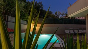 A swimming pool with a tiled deck, surrounded by green plants and a fence.
