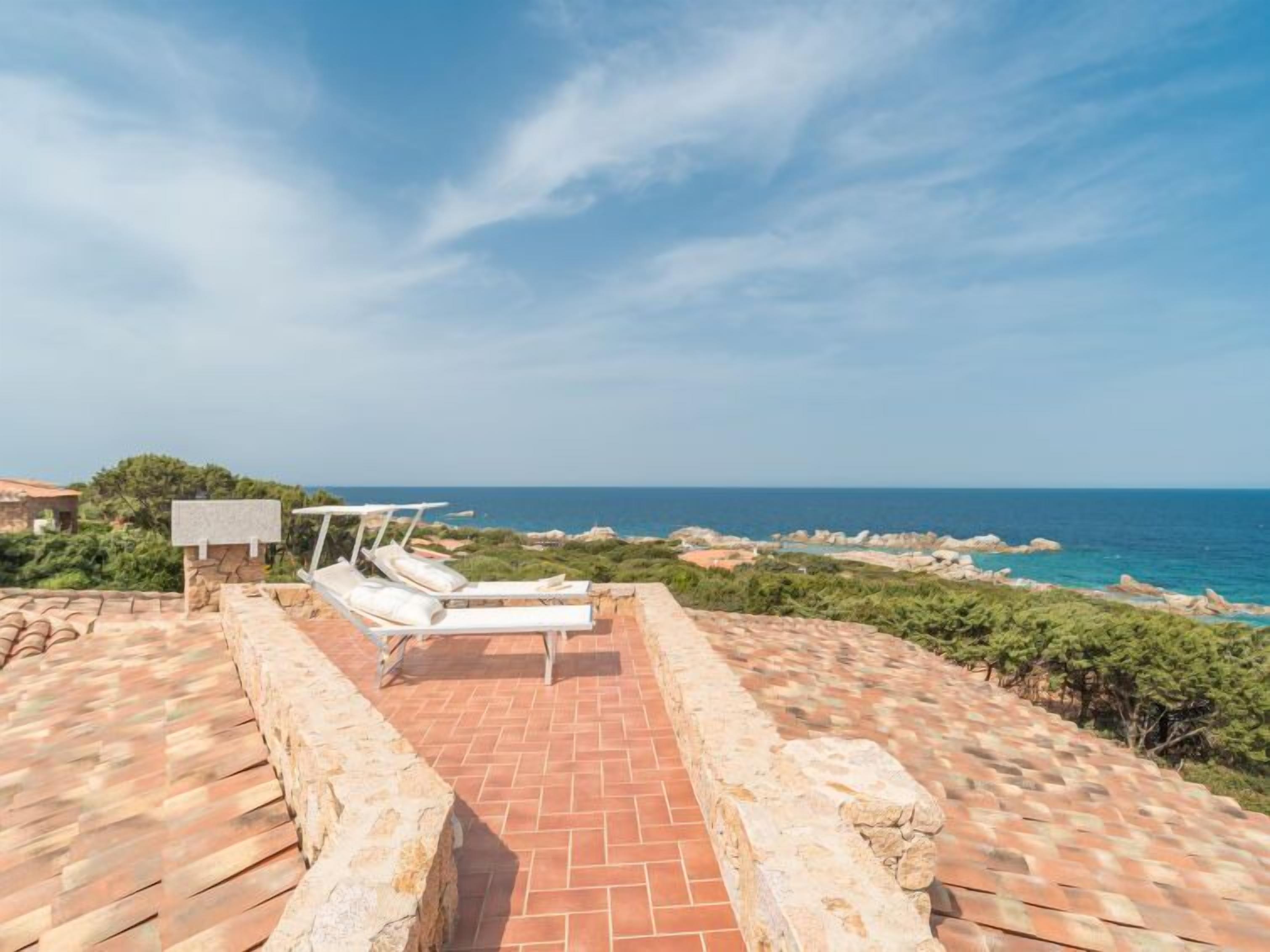 A rooftop terrace with a stone pathway leading to a seating area with two white lounge chairs and a small table, overlooking a coastal landscape with rocky outcrops and the sea.