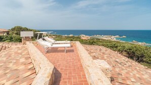 Eine Dachterrasse mit einem Steinweg, der zu einer Sitzgelegenheit mit zwei weißen Liegestühlen und einem kleinen Tisch führt, mit Blick auf eine Küstenlandschaft mit Felsformationen und dem Meer.
