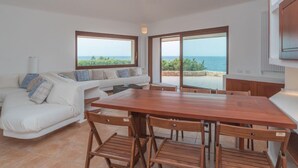 Ein modernes Wohnzimmer mit einem Holzesstisch, einer weißen Couch und großen Fenstern mit Blick auf das Meer.