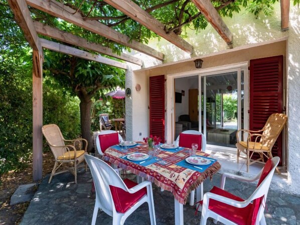Eine Terrasse im Freien mit einem Tisch für vier Personen, umgeben von weißen Stühlen mit roten Kissen, unter einer Holzpergola.