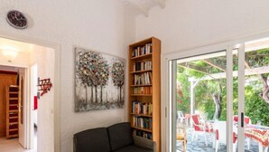 A living room with a dark grey sofa, a bookshelf, a painting of trees, and a sliding glass door leading to an outdoor patio with chairs and tables. - Villa Irène by Interhome (Bravone)