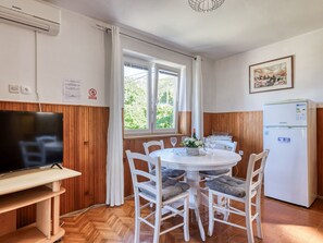 A dining area with a round table, white chairs, a television on a stand, and a refrigerator.