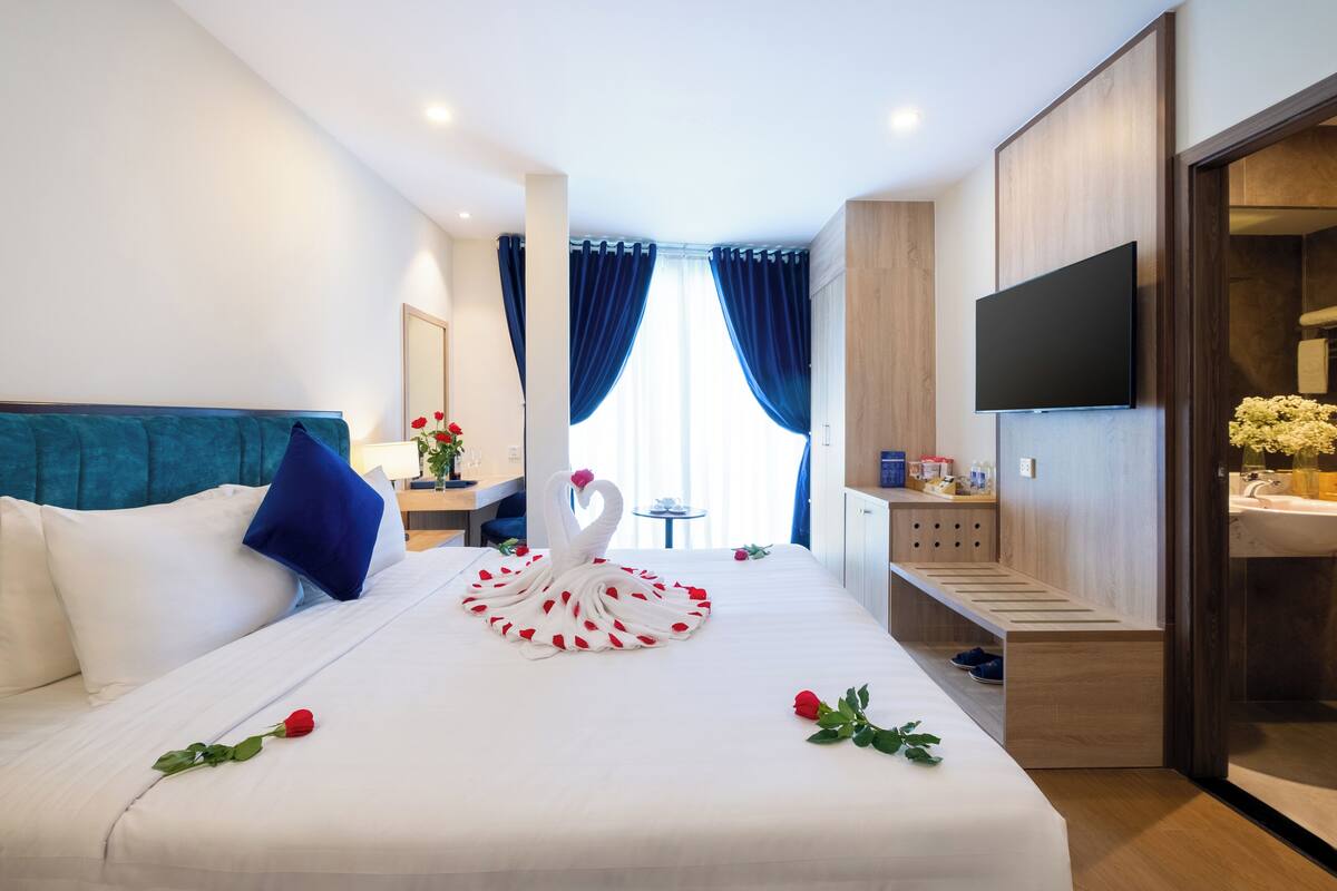 A hotel room with a neatly made bed, a TV mounted on the wall, a desk with a chair, and a bathroom visible through an open door.