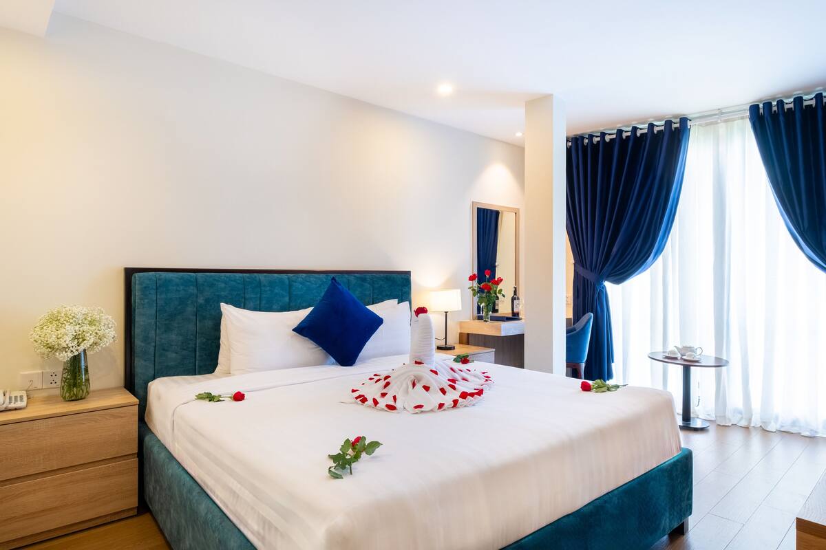 A hotel room with a large bed, a bedside table with a vase of flowers, a nightstand, a mirror, a small table with a lamp, and a view of the outdoors through large windows with curtains.