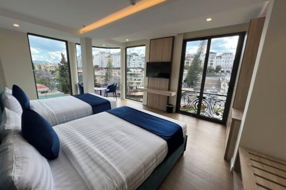 Una habitación de hotel moderna con dos camas, televisor y ventanas amplias que ofrecen vistas a la ciudad.