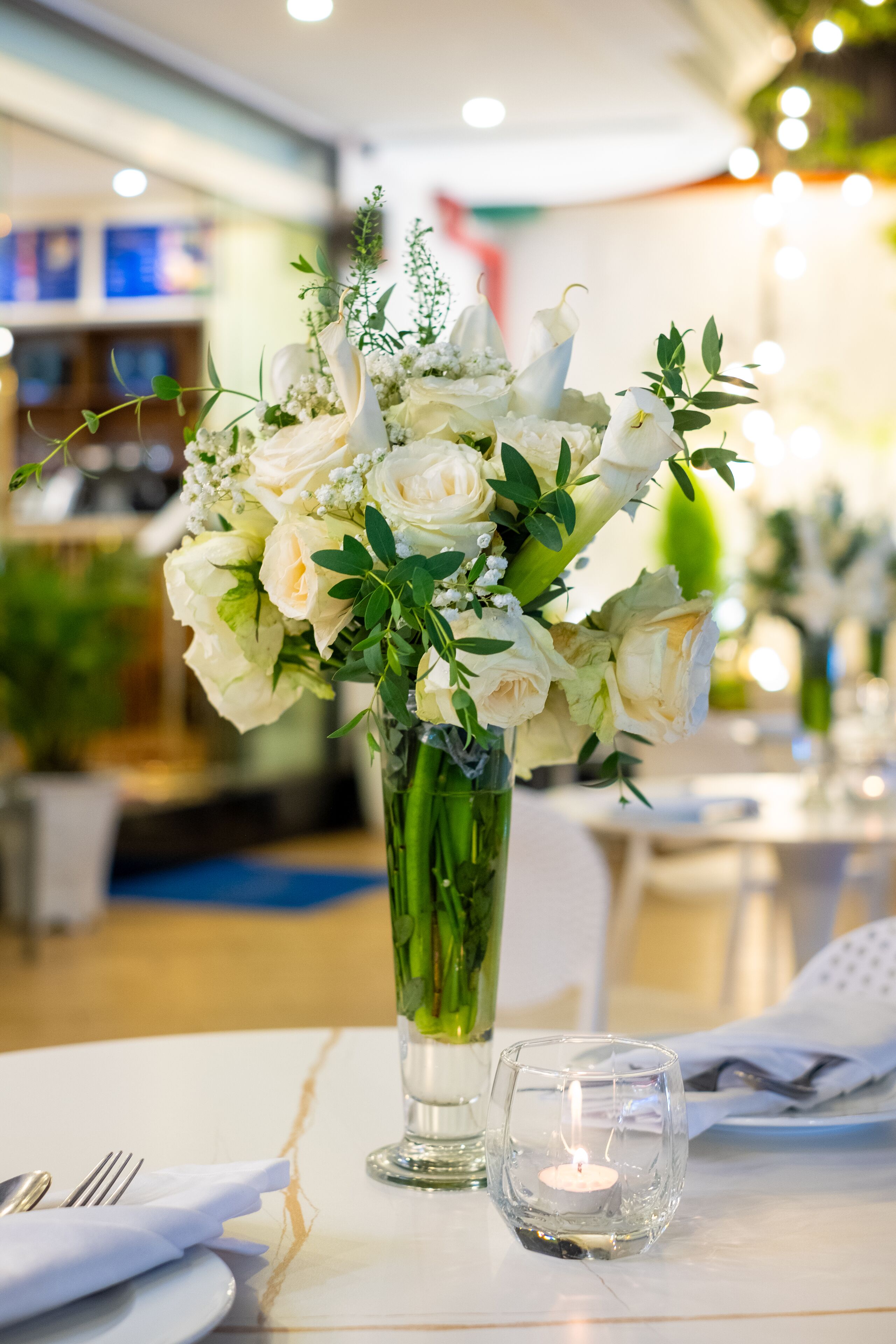 Una mesa con un florero de flores blancas, una vela encendida y una servilleta doblada.