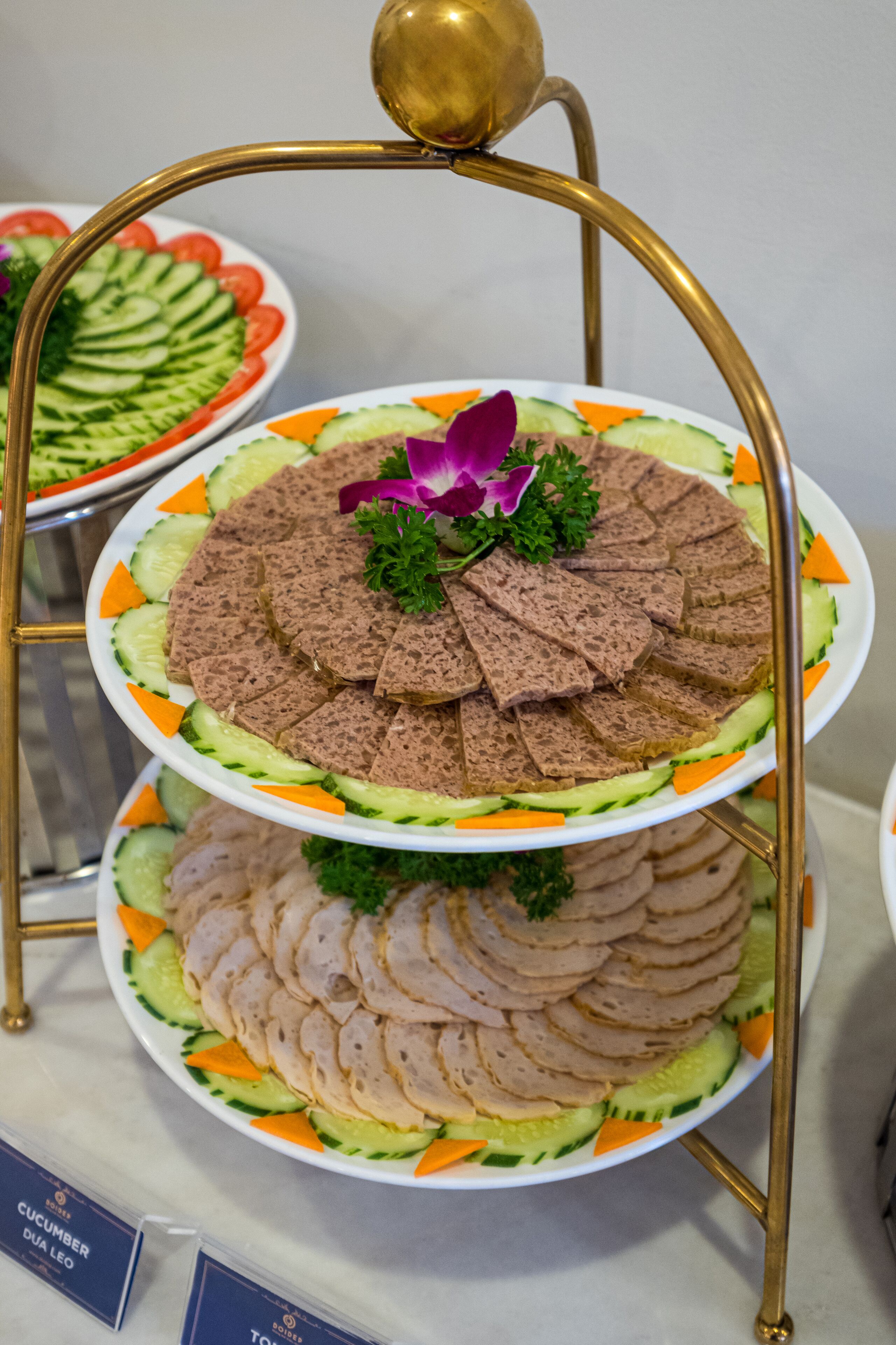 A multi-tiered platter with sliced meats and vegetables, displayed on a gold stand.