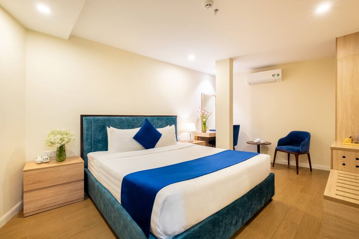 A modern hotel room with a large bed, a bedside table, a chair, a desk, and an air conditioning unit.
