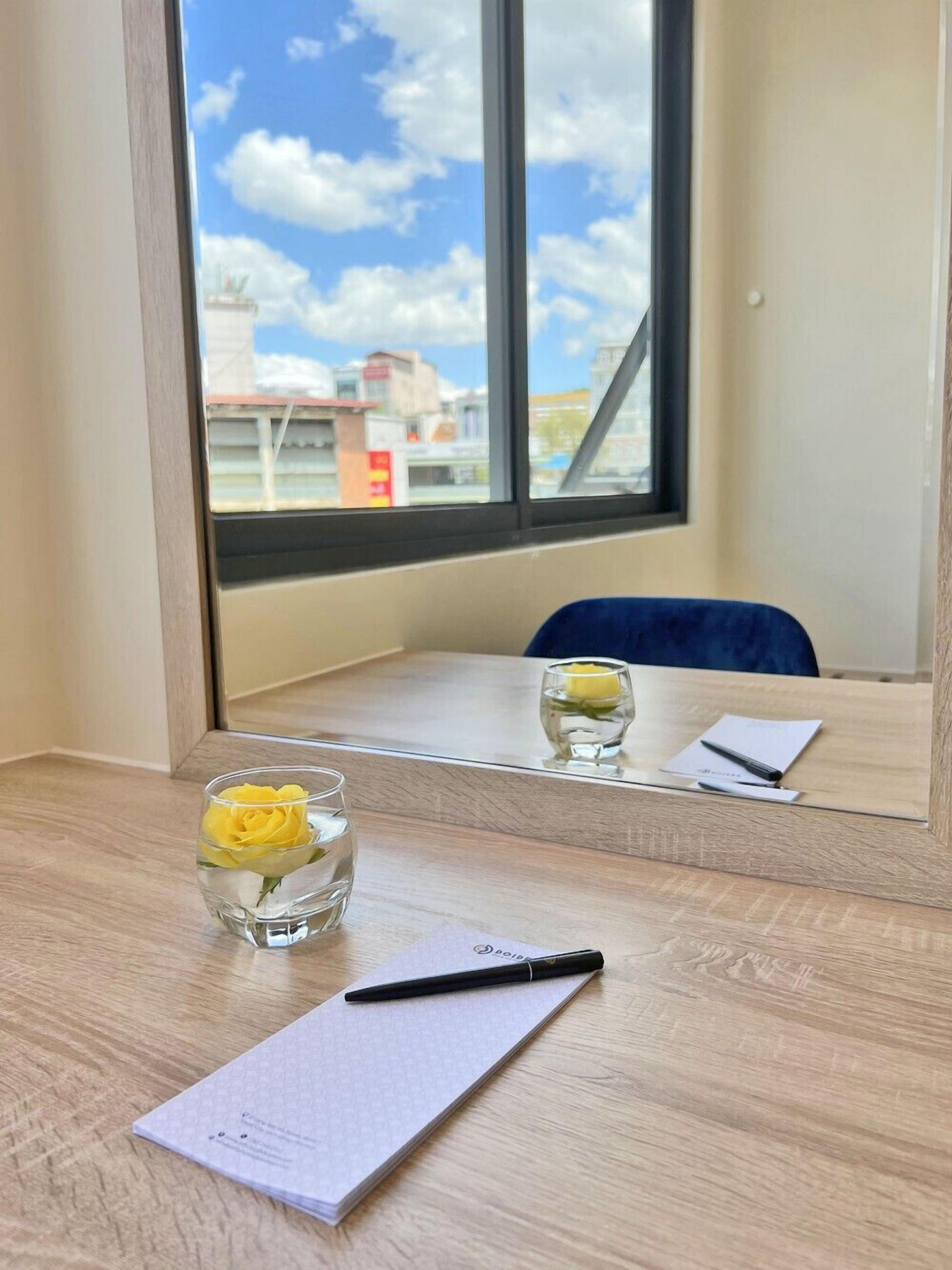 Habitación de hotel con un escritorio de madera, un cuaderno, un bolígrafo y un vaso de agua con una flor amarilla.