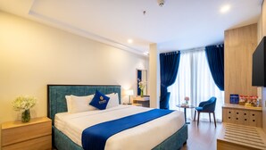 A hotel room with a large bed, a bedside table with a vase of flowers, a small table with a lamp, a chair, and a TV on a wooden cabinet. - Sandals Flora Hotel (Xuan Huong - Da Lat)