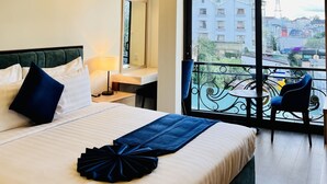 Habitación de hotel con cama, mesita de noche, lámpara y un balcón con vistas a la ciudad.
