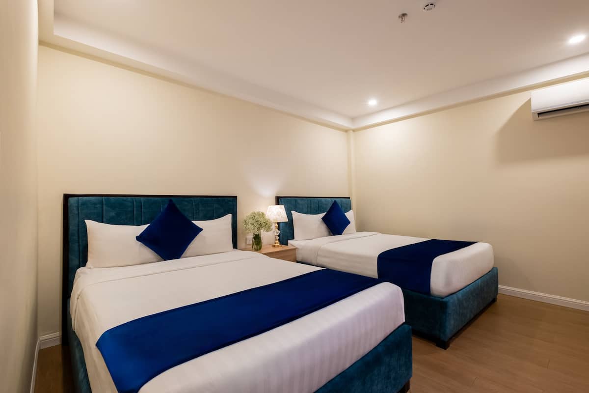 A hotel room with two beds, each with a blue headboard and white bedding. There is a nightstand with a lamp and a small plant between the beds.