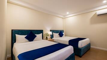 Quarto de hotel com duas camas, cada uma com cabeceira azul e roupa de cama branca. Há uma mesinha de cabeceira com uma luminária e uma planta pequena entre as camas.