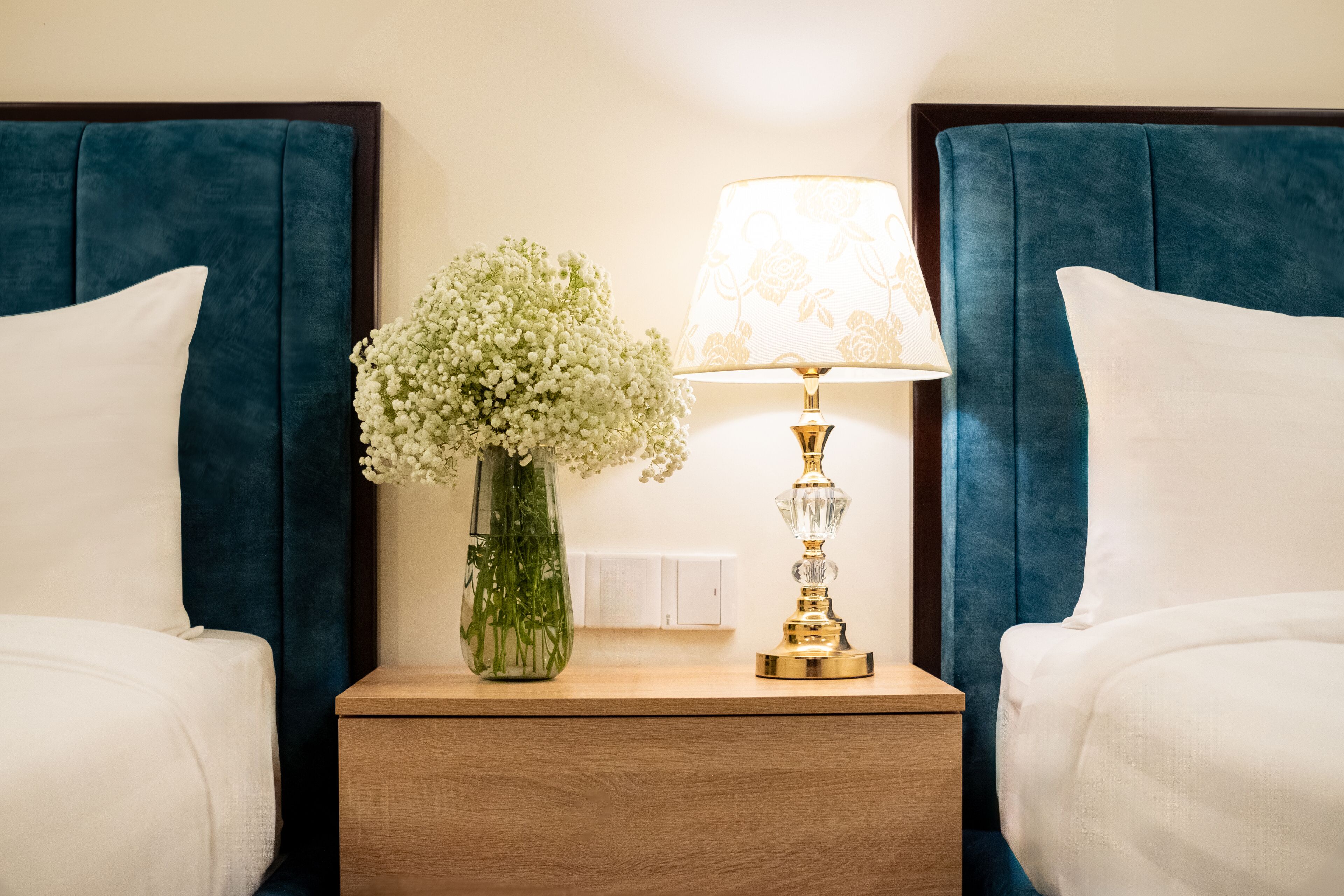 Una cama bien tendida con sábanas blancas, una mesita de noche con un jarrón de flores blancas y una lámpara con pantalla floral.