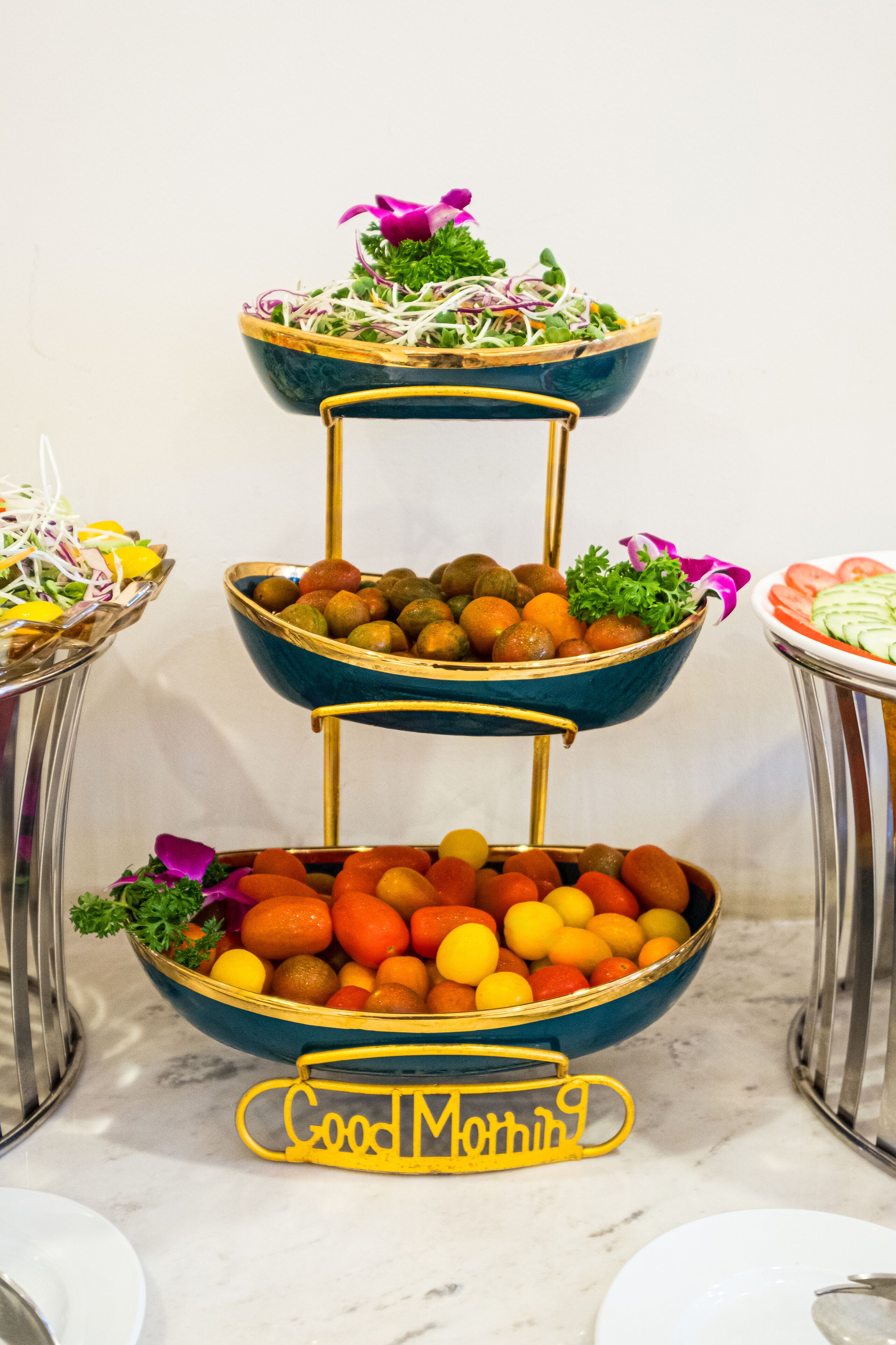 A tiered serving tray with various fruits and vegetables, including oranges and greens, on a marble countertop.