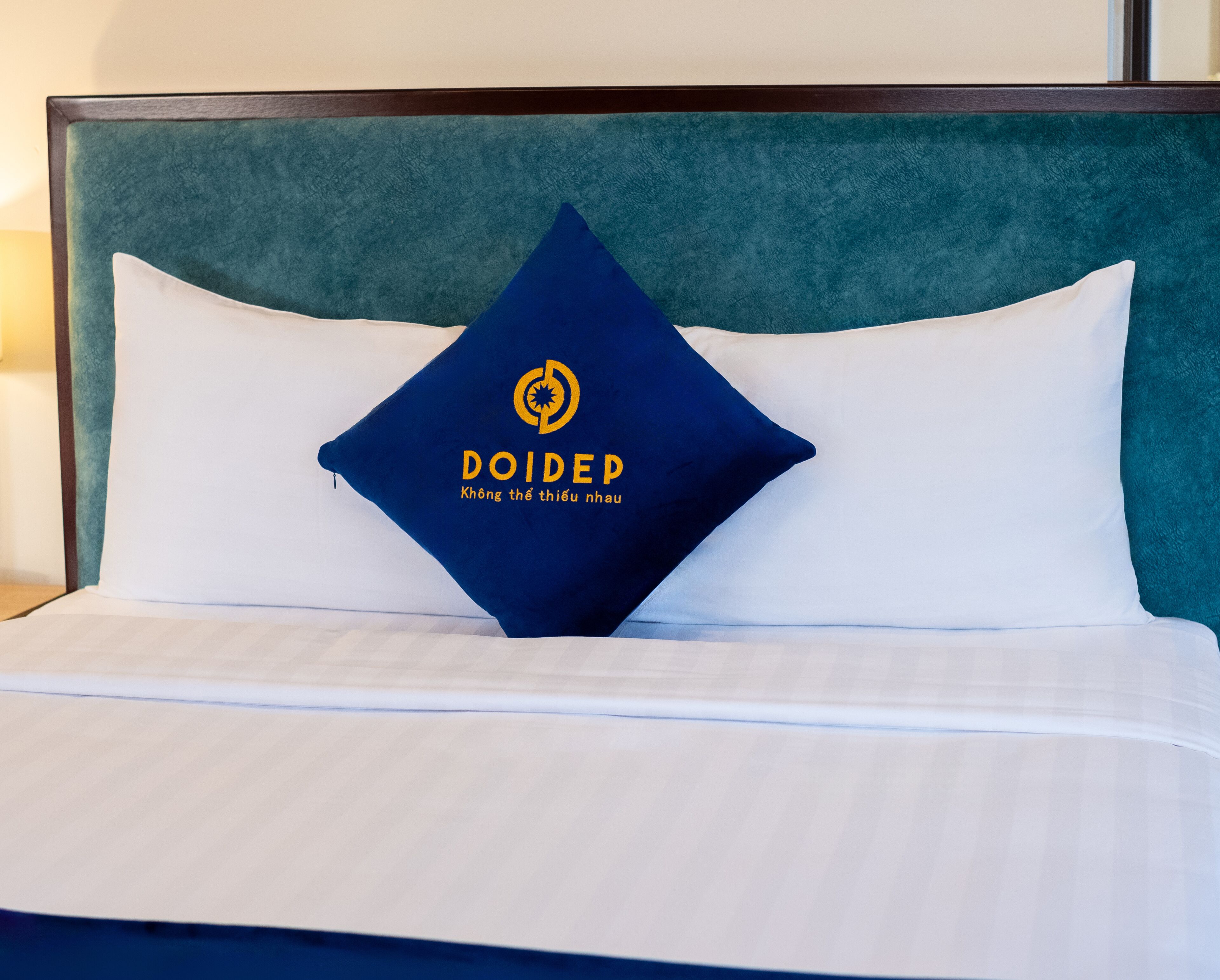 A bed with white bedding and a teal headboard. A blue pillow with a logo and text is placed on the bed.