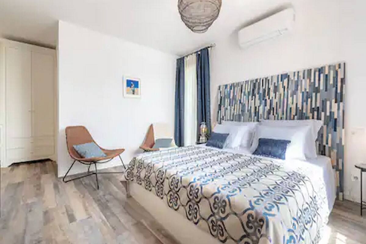 Un dormitorio moderno con una cama grande, dos sillas, una lámpara colgante y una obra de arte en la pared.