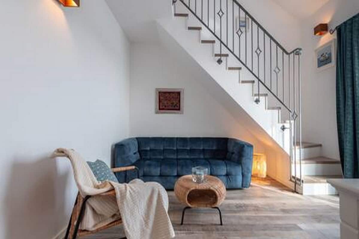 Una sala de estar moderna con un sofá azul, una silla de madera, una mesa de centro de vidrio y una escalera con barandilla de metal.