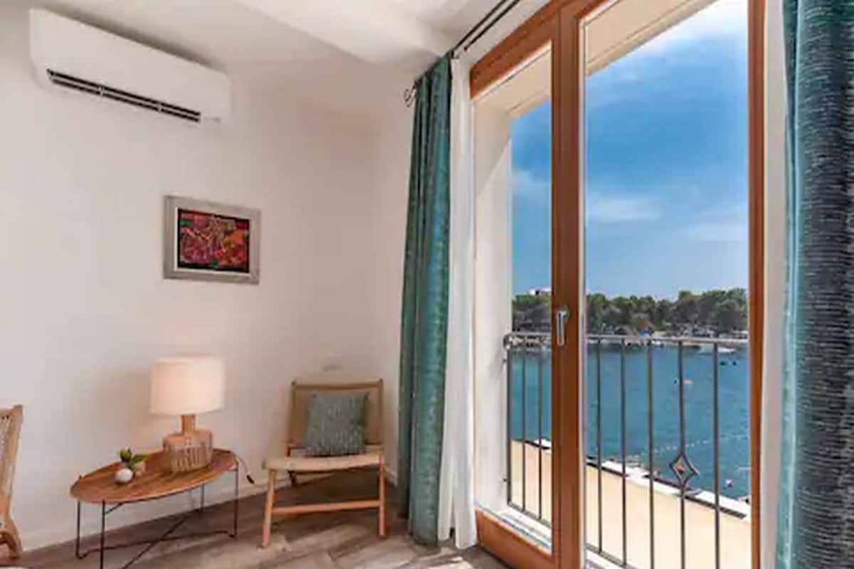 Una habitación con piso de madera, una silla, una mesa con lámpara, un cuadro en la pared y un amplio ventanal con vista al mar y un balcón.
