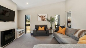A modern living room with a fireplace, a grey sofa with orange cushions, a black armchair, and a large TV mounted on the wall. - If I can dream with wifi & pets, room to park the boat (Taupō)