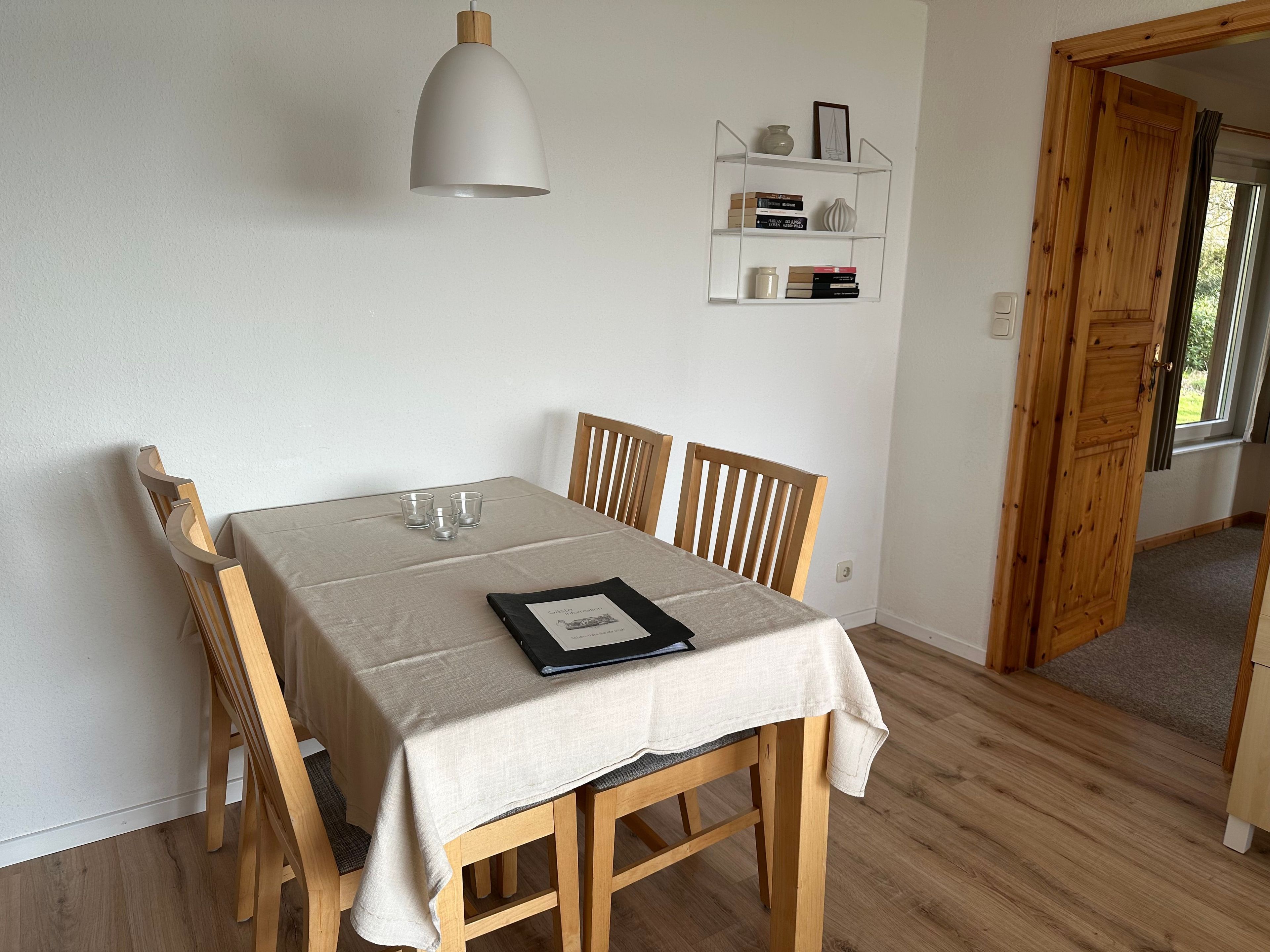 A dining table with a white tablecloth, wooden chairs, and a pendant light. A bookshelf with books and decorative items is mounted on the wall.