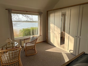 A room with a large window overlooking a body of water, wicker chairs, a small table, and a wardrobe with sliding doors.