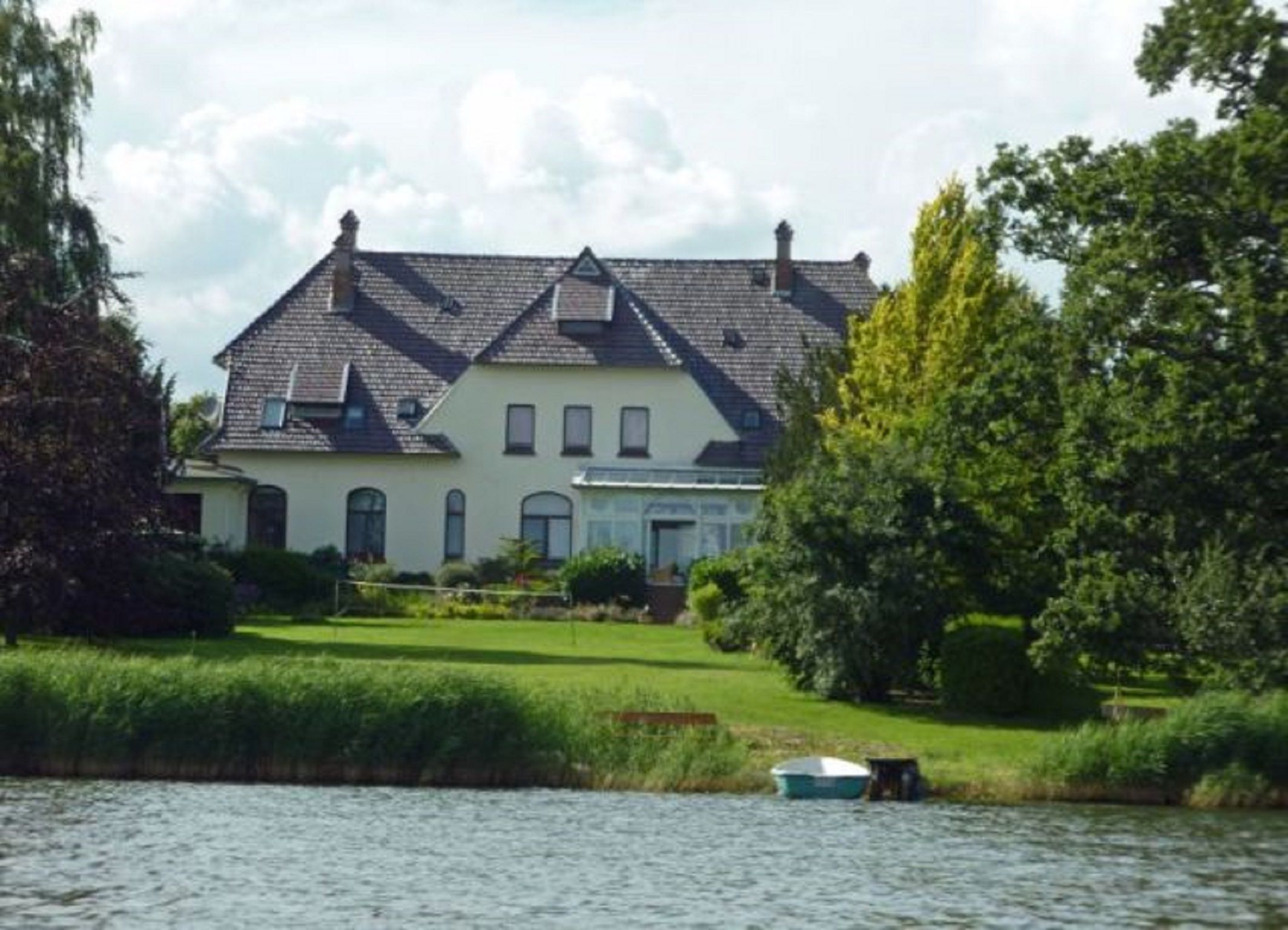 A large house with a sloped roof, a front porch, and a small boat on the water in front.