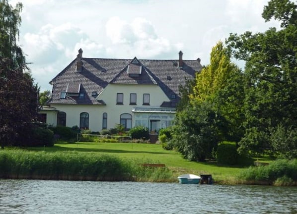 A large house with a sloped roof, a front porch, and a small boat on the water in front.