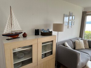 A modern living room with a wooden cabinet containing glassware, a model sailboat, and a vase. A grey sofa and a floor lamp are visible, with a window offering a view of greenery.