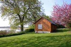 A wooden cabin with a small window, surrounded by lush greenery and a tree with pink blossoms.