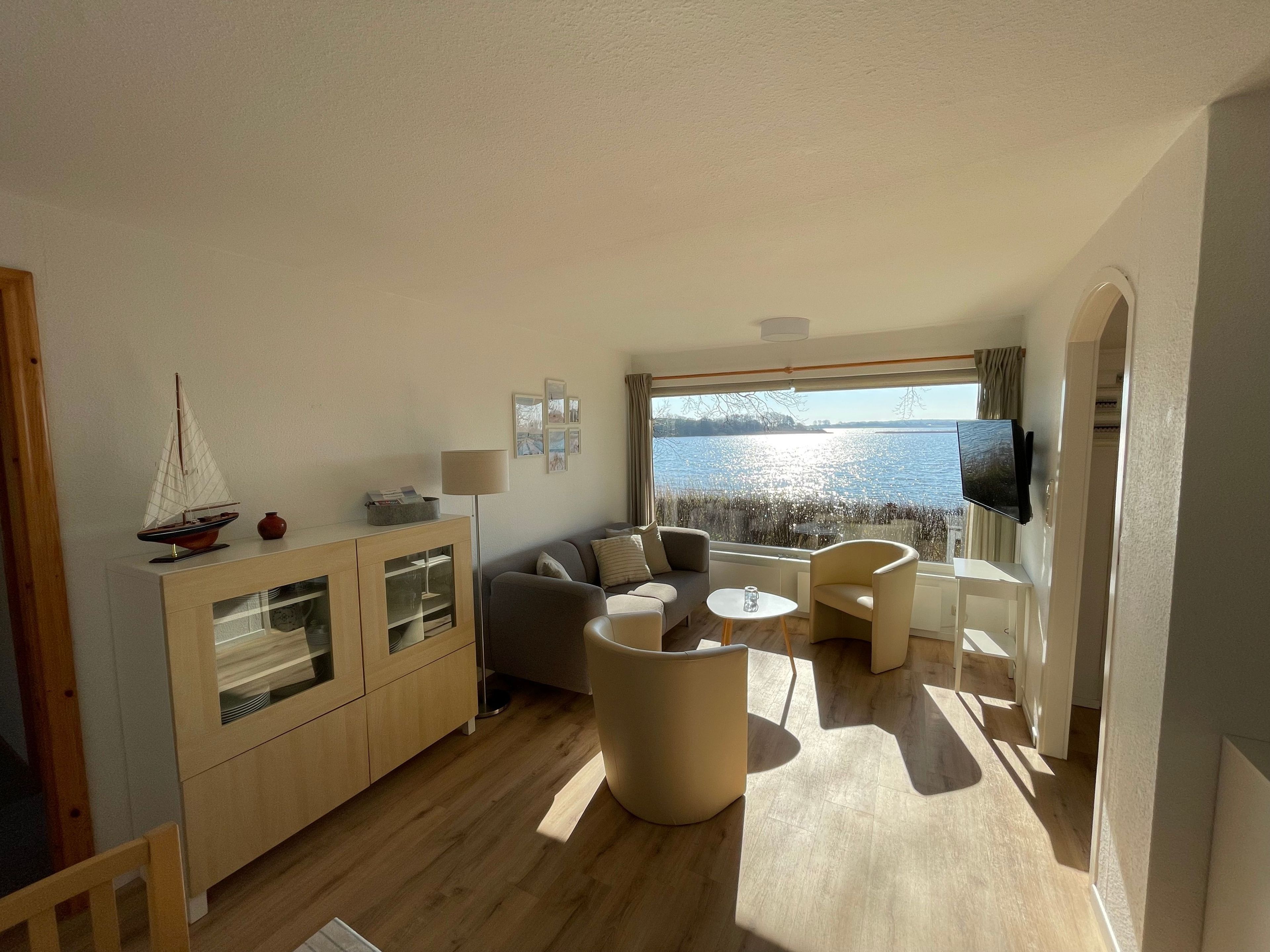 A modern living room with a view of a lake, a wooden cabinet with glass doors, a sofa, a coffee table, and a TV mounted on the wall.