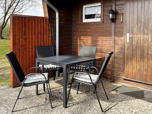 En uteplats med ett bord och fyra stolar, en trävägg, ett fönster och en väggmonterad lampa.