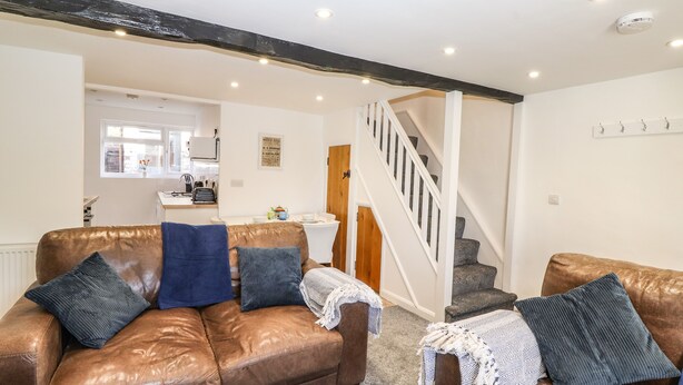 A cozy living room with leather sofas, a staircase, and a kitchen in the background. - Sparrow Cottage (Keswick)