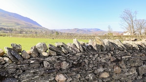 A stone wall with sheep grazing in a green field, mountains in the background, and a clear blue sky. - Sparrow Cottage (Keswick)