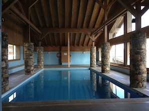 Piscina interior com pilares de pedra, vigas de madeira e amplas janelas.