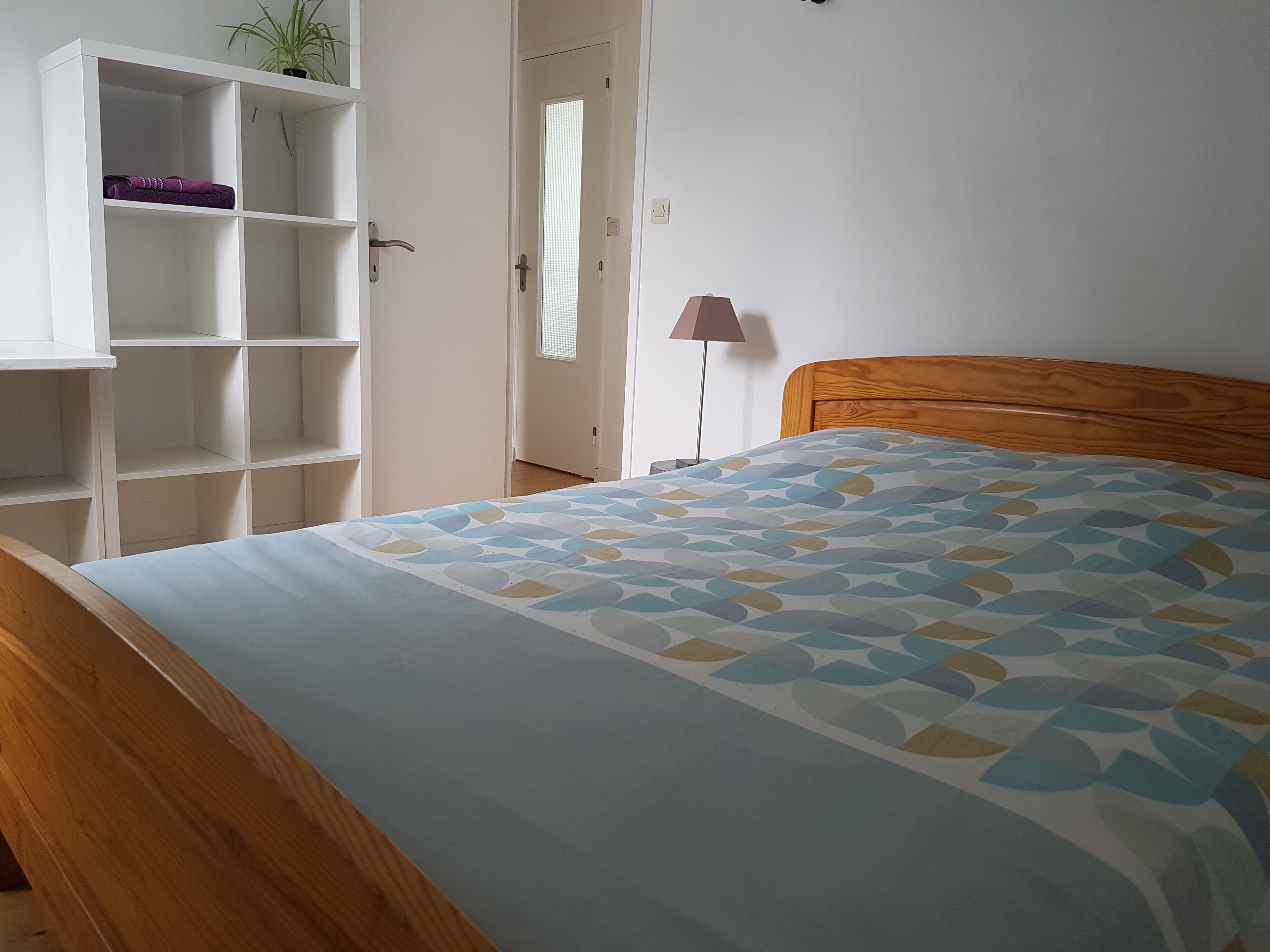 Een slaapkamer met een houten hoofdbord, een bed met een geborduurde spreideken, een witte garderobe met planken, en een potplant op de bovenste plank.