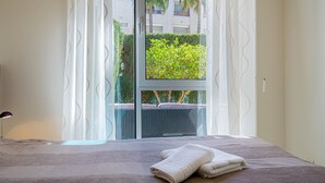 A bed with a folded towel, a window with a view of greenery, and a lamp on a bedside table. - Apartment 'Estrella 2 3 BH' close to the Beach with Shared Pool & Wi-Fi (l'Alfàs del Pi)