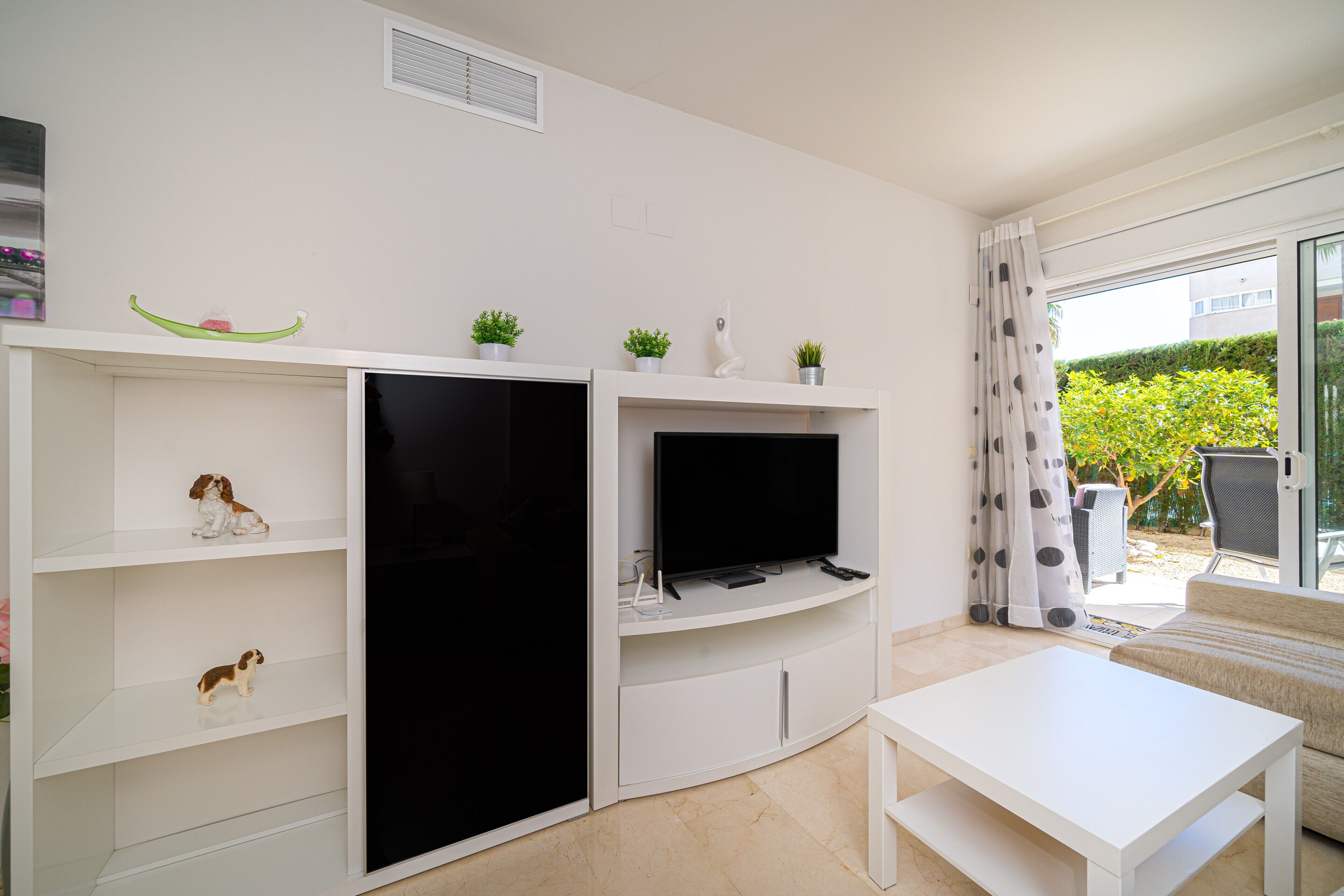 A modern living room with a flat-screen TV, white shelves, a small sofa, and a dining table.
