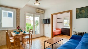 A living room with a blue sofa, a wooden dining table with chairs, a TV mounted on the wall, and a balcony with a view of buildings and greenery. - Pet-Friendly Apartament Luisa close to the Beach with Balcony & Wi-Fi (Lloret de Mar)