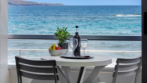 Una mesa con una botella, vasos y un cuenco de fruta en un balcón con vista al mar.