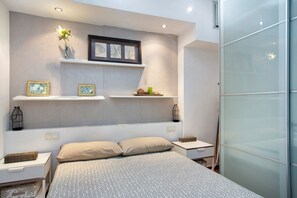 A modern bedroom with a bed, bedside tables, a wall-mounted shelf with decorative items, and a frosted glass partition.
