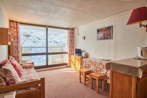 Een knus ingerichte kamer met een bed, een klein tafeltje, een bureau, een stoel, een dressoir en een raam met uitzicht op besneeuwde bergen.