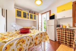 Une cuisine avec une table à manger, des chaises, un saladier rouge, un ventilateur et un réfrigérateur.