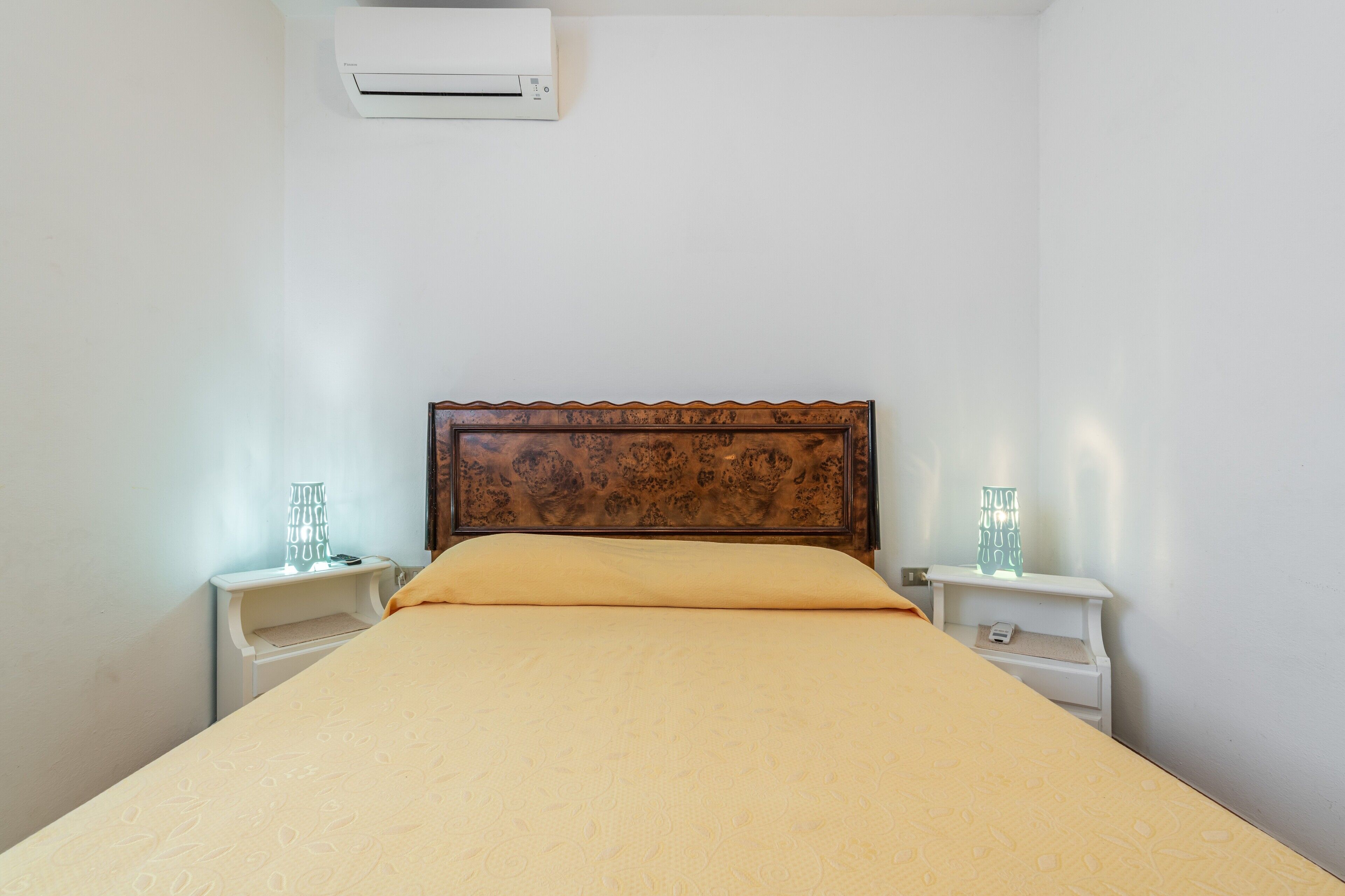 Una camera da letto con una testiera in legno, un letto con copripiumino color beige, due comodini con lampade e un condizionatore d'aria a parete.